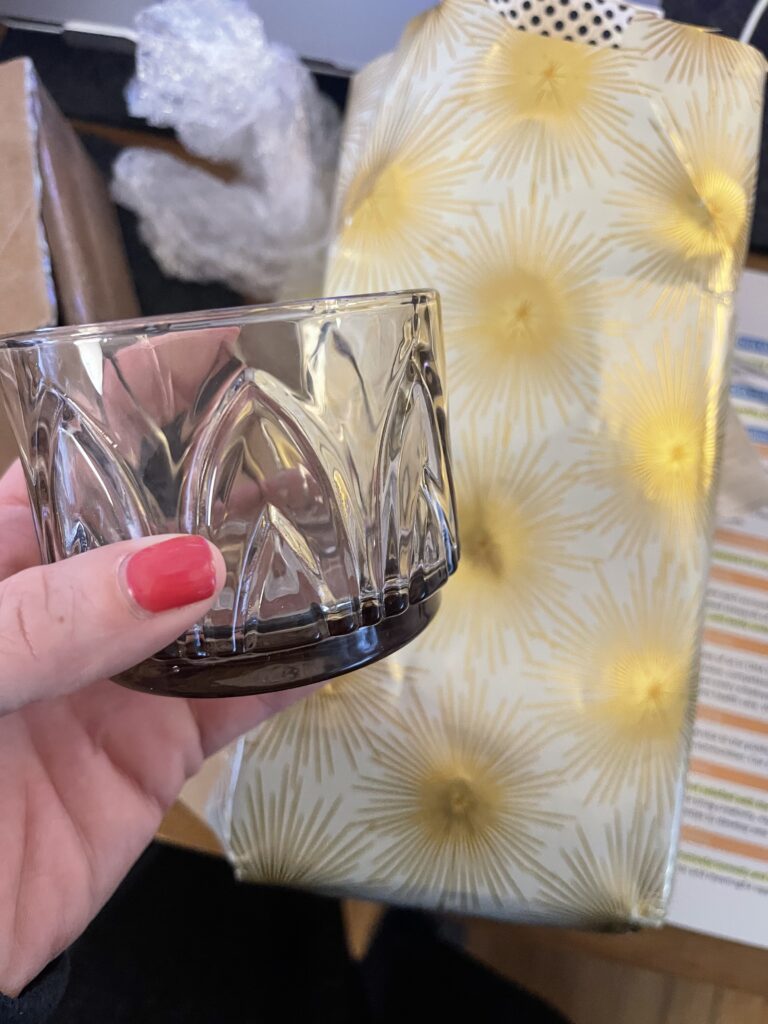 My disembodied hand holds a small decorative glass unexpectedly gifted to me by my former roommate. Shiny gold wrapping paper is in the background. 