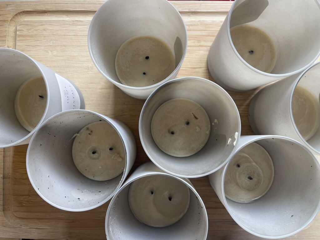 An overhead view of eight candles of the same scent burned down to the bottom. The candles are on a wooden cutting board waiting for me to place them one or two at a time into a pot of boiling water to melt down the wax so I can pour it into the decorative glass gifted to me by my former roommate to make a candle. 