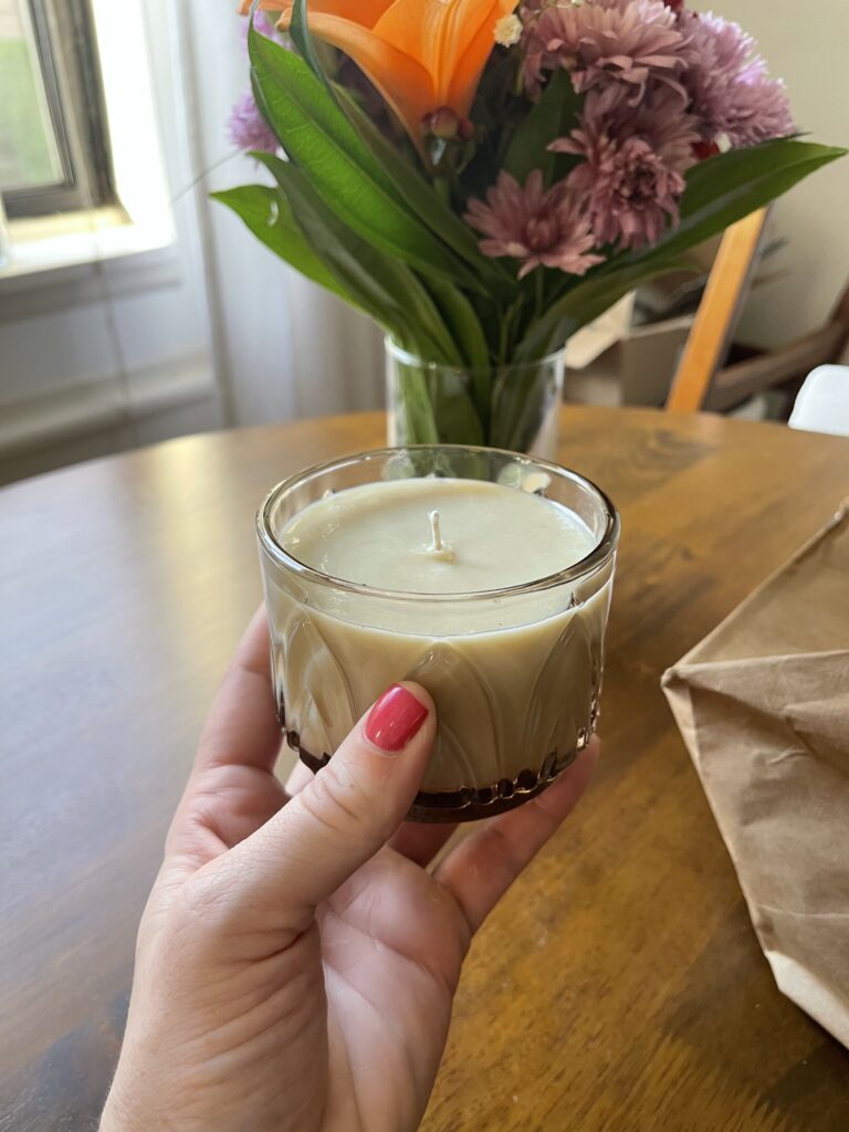 My disembodied hand holds against a wood table with a vase full of flowers on it a small candle that I made using the leftover wax from eight candles (of the same scent) and a decorative glass gifted to me by my former roommate. 