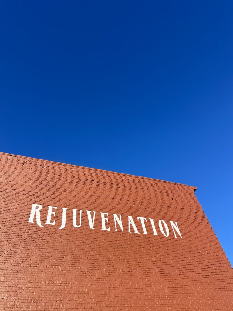 A bright red brick wall of the exterior of a building reads "REJUVENTATION" in white paint against a clear, very blue sky. 