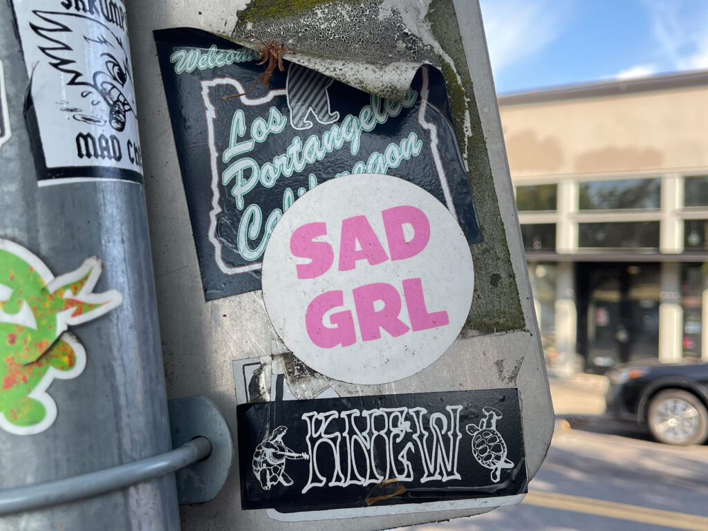 The back of a street sign in Portland, Oregon, with a white circular sticker that says "SAD GRL" in bubblegum pink. 