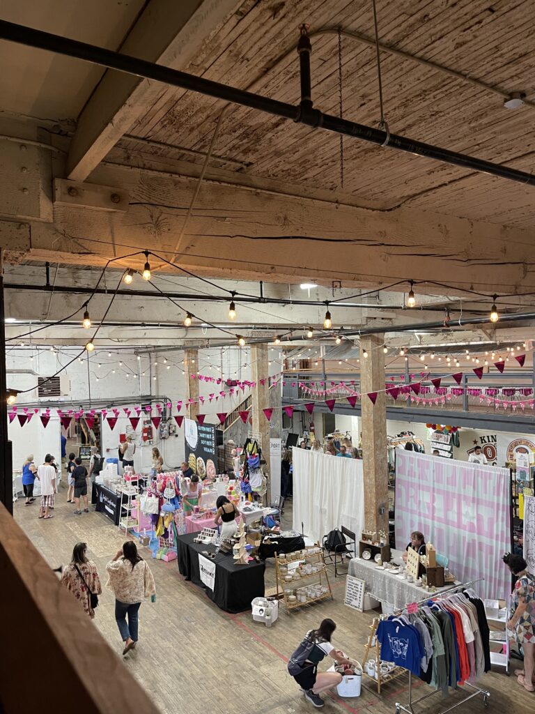 A view of the first few minutes of Portland Night Market in July 2024, as seen from above. A handful of tables with various goods such as clothing, jewelry, and baked goods are set up in the space and a few people are walking around. 