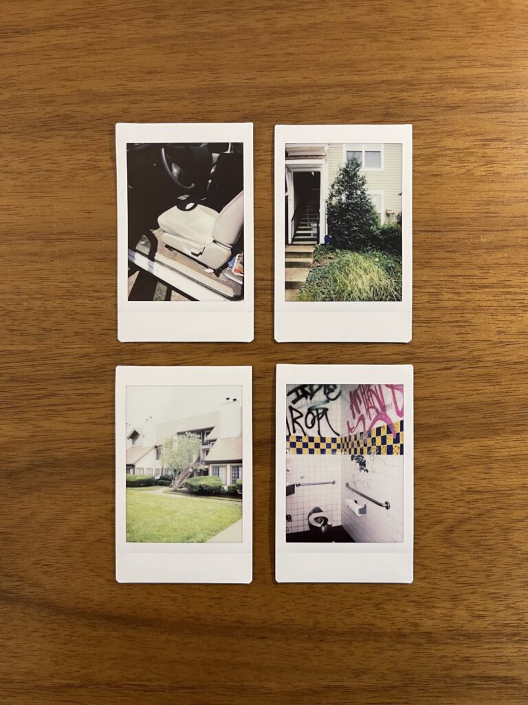 Four Instax/Polaroid photos face up on a wooden desk. They're arranged in two rows of two photos each. Two photos depict the exterior of different apartment buildings. One photo depicts the inside cap of a pickup truck. The final photo depicts the inside of a public restroom. 