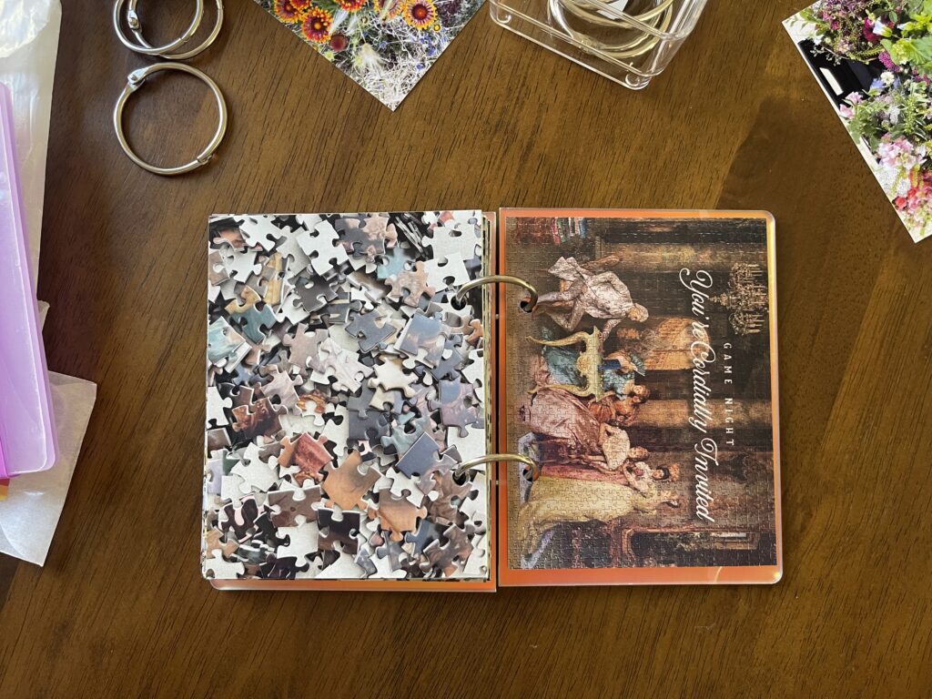 Spread #21. The left-hand page is a photo of the puzzle's unpuzzled pieces. The right-hand page is a photo of the completed puzzle of an old painting of wealthy women and one man in 1800s-style clothing seated at a table playing Connect Four and surrounded by other modern-day games, like Uno and Twister. 